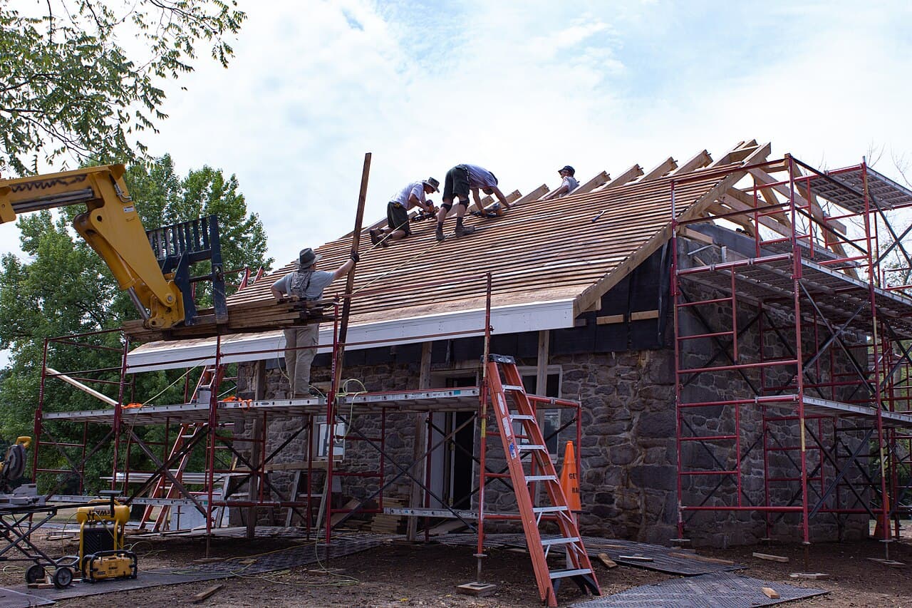 Roofing crew installing new cedar shingles on a historic stone house with scaffolding around it